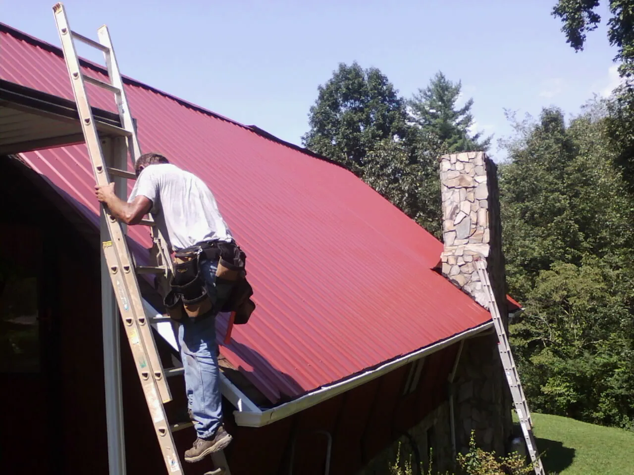 Roofer on a ladder beside a red metal roof and stone chimney — Soffit & Fascia Repair by Roofing Brothers in Garibaldi